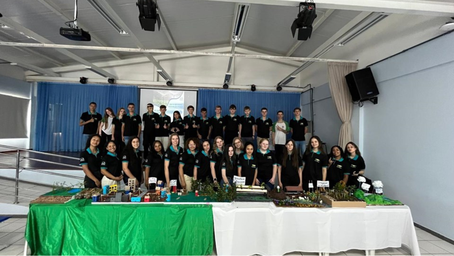 Alunos apresentando maquetes coloridas que representam sistemas de irrigação e ciclos da água, com professores e convidados observando atentamente.